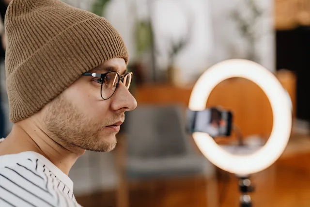 A man wearing eyeglasses and a knitted cap records a video at home using a smartphone and ring light. Balancing exclusivity with a creator's existing partnerships