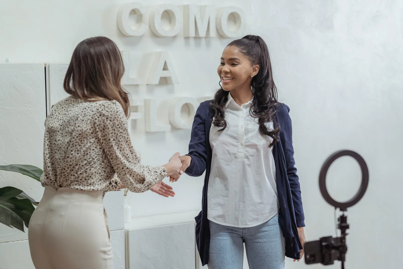 Positive African American female shaking hand of partner standing in light office near smartphone with ring lamp on tripod