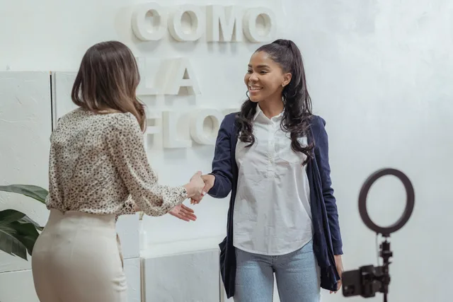 Positive African American female shaking hand of partner standing in light office near smartphone with ring lamp on tripod. Building an influencer ambassador programme