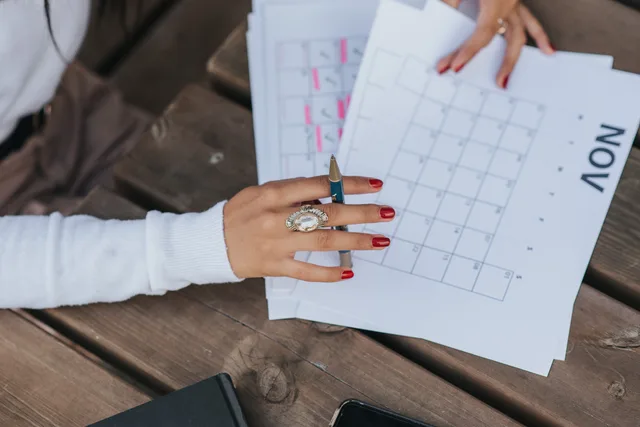 From above crop anonymous female in white blouse sitting at wooden table with pen and making list of plans for month. Planning ambassador deliverables across a quarter